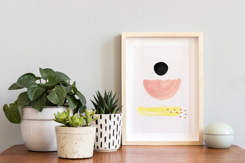 May include: A framed abstract print with a black circle, pink half-circle, and yellow stripe on a white background. Three potted plants are on a wooden surface in front of the print.