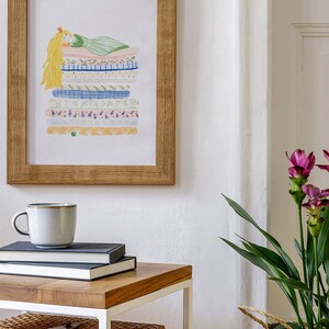 May include: A framed illustration of a princess lying on a stack of colorful patterned fabric. The illustration is in a light wood frame and is hanging on a white wall. A white side table with a wooden top is in the foreground with a mug, books, and two wicker baskets.