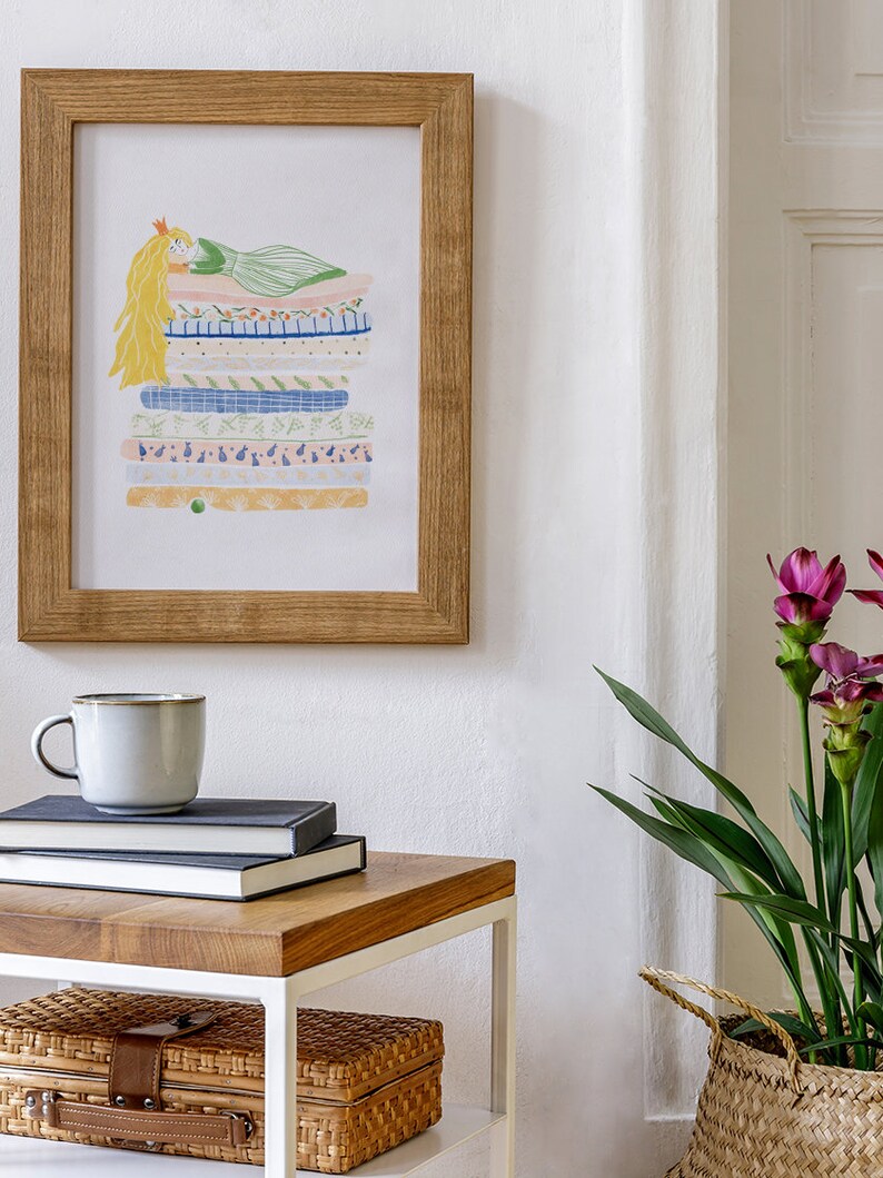 May include: A framed illustration of a princess lying on a stack of colorful patterned fabric. The illustration is in a light wood frame and is hanging on a white wall. A white side table with a wooden top is in the foreground with a mug, books, and two wicker baskets.