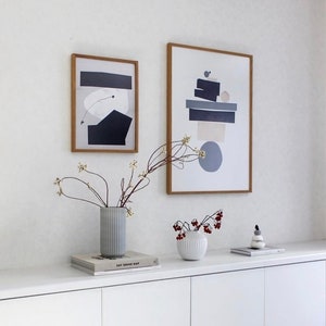 May include: Two framed abstract art prints with geometric shapes in shades of gray, blue, and beige, hanging on a white wall above a white dresser with two vases and a book titled "Eat Drink Nap".
