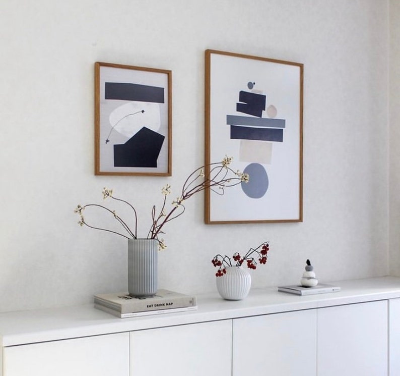May include: Two framed abstract art prints with geometric shapes in shades of gray, blue, and beige, hanging on a white wall above a white dresser with two vases and a book titled "Eat Drink Nap".