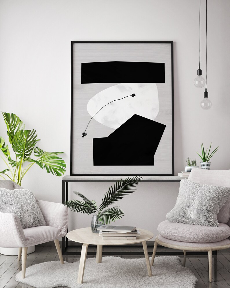 May include: A black and white abstract art print in a black frame hangs above a white console table with a potted plant and two armchairs with fluffy white pillows. The room has a white wall and a light wood floor.