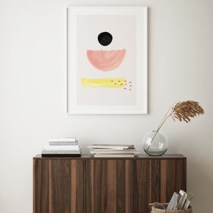 May include: A framed abstract print with a black circle, a pink half-circle, and a yellow rectangle with red dots. The print is hanging on a white wall above a wooden cabinet with a vase and a basket.