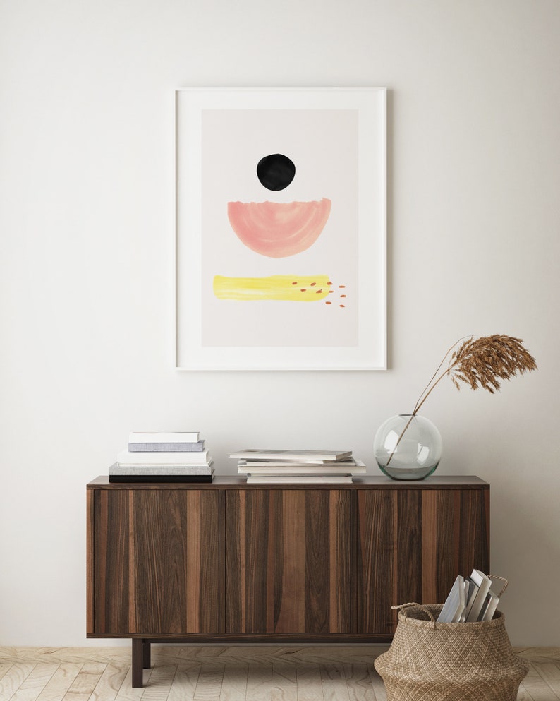 May include: A framed abstract print with a black circle, a pink half-circle, and a yellow rectangle with red dots. The print is hanging on a white wall above a wooden cabinet with a vase and a basket.