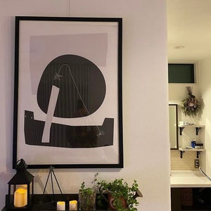 Puede incluir: Una impresi&oacute;n enmarcada en negro con fondo blanco y formas abstractas en blanco y negro. La impresi&oacute;n est&aacute; colgada en una pared blanca sobre una estanter&iacute;a con velas y plantas.