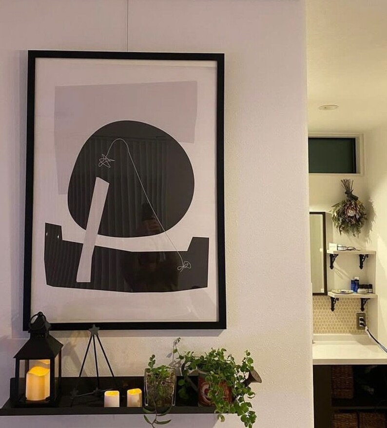 Puede incluir: Una impresi&oacute;n enmarcada en negro con fondo blanco y formas abstractas en blanco y negro. La impresi&oacute;n est&aacute; colgada en una pared blanca sobre una estanter&iacute;a con velas y plantas.
