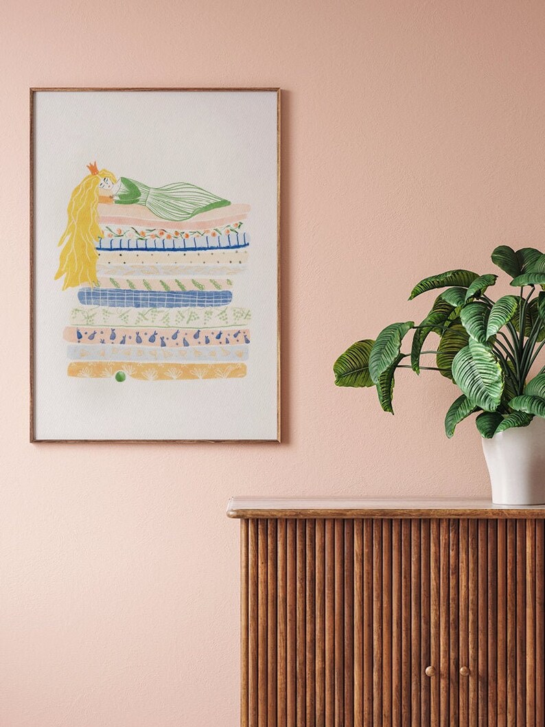 May include: A framed illustration of a princess with long blonde hair lying on a stack of colorful patterned mattresses. The princess is wearing a green dress and a gold crown. The illustration is on a light pink wall above a wooden cabinet.