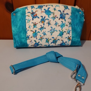 May include: A white and blue fabric pouch with a zipper closure and a turquoise blue strap. The pouch has a pattern of colorful sea turtles.