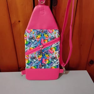 May include: A pink and blue crossbody bag with a zipper closure and a pattern of cartoon characters. The bag has a pink strap and a silver buckle.