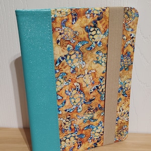 May include: A teal blue notebook cover with a brown elastic closure. The cover features a pattern of colorful sea turtles on a yellow background.