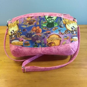 May include: A pink fabric handbag with a zipper closure and a long pink strap. The bag has a colorful print featuring Harry Potter themed images, including the Hogwarts houses, characters, and magical objects.