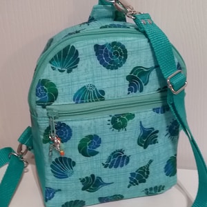 以下が含まれることがあります： 貝殻柄のターコイズブルーのクロスボディバッグ。バッグにはジッパー付きのコンパートメントが2つと、調節可能な長いストラップが付いています。