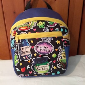 May include: A small blue backpack with a black handle and yellow trim. The front of the backpack is covered in a black and white print of potion bottles, hearts, stars, and the text "Sleeping Draught", "Draught of Peace", "Sleeheazy's Hair Potion", and "Half Pot".