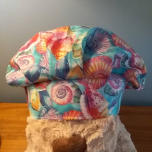 May include: A turquoise blue chef hat with a colorful seashell pattern. The hat is made of fabric and has a white brim.