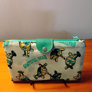 May include: A light blue rectangular fabric wallet featuring a repeating pattern of turtles playing musical instruments and the text Grateful Dead in teal. The wallet includes a teal snap-button closure flap and a matching teal zipper along the top edge.
