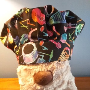 Può includere: Un cappello da chef nero con una stampa colorata di strumenti musicali. La stampa presenta chitarre, batterie, trombe e altri strumenti musicali.