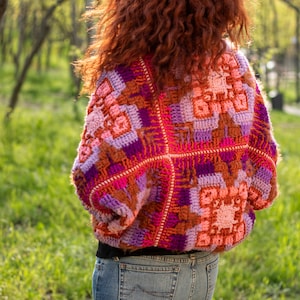 May include: A woman wearing a colorful crocheted sweater with a geometric pattern in shades of pink, purple, orange, and brown. The sweater has a loose fit and is worn over a black shirt and blue jeans.