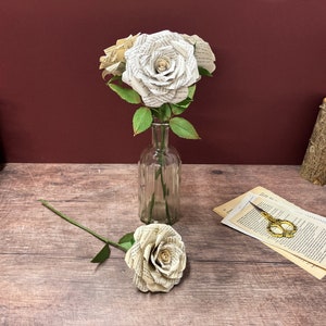 May include: Three white paper roses made from book pages in a clear glass vase on a wooden surface. A single white paper rose with a green stem is lying on the surface.