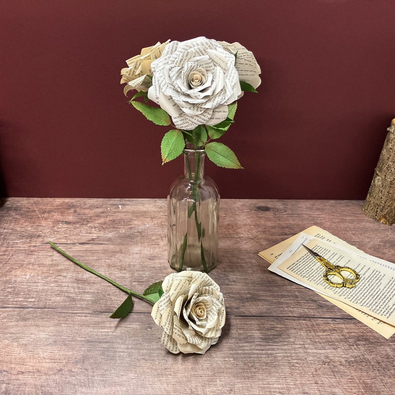 May include: Three white paper roses made from book pages in a clear glass vase on a wooden surface. A single white paper rose with a green stem is lying on the surface.