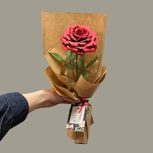 May include: A single, large red rose with green leaves, wrapped in brown paper. The rose is tied with a red ribbon and a tag that reads "A Long's Story Gifts for Readers". The rose is held by a person.