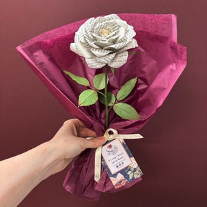 May include: A white paper rose made from book pages, with green leaves and a brown stem, is wrapped in burgundy tissue paper and tied with a white ribbon. A tag with the text "A Long's Story Gift for Readers" is attached to the ribbon.