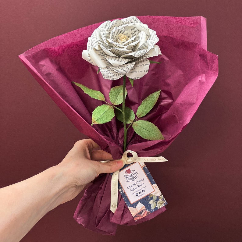 May include: A white paper rose made from book pages, with green leaves and a brown stem, is wrapped in burgundy tissue paper and tied with a white ribbon. A tag with the text "A Long's Story Gift for Readers" is attached to the ribbon.
