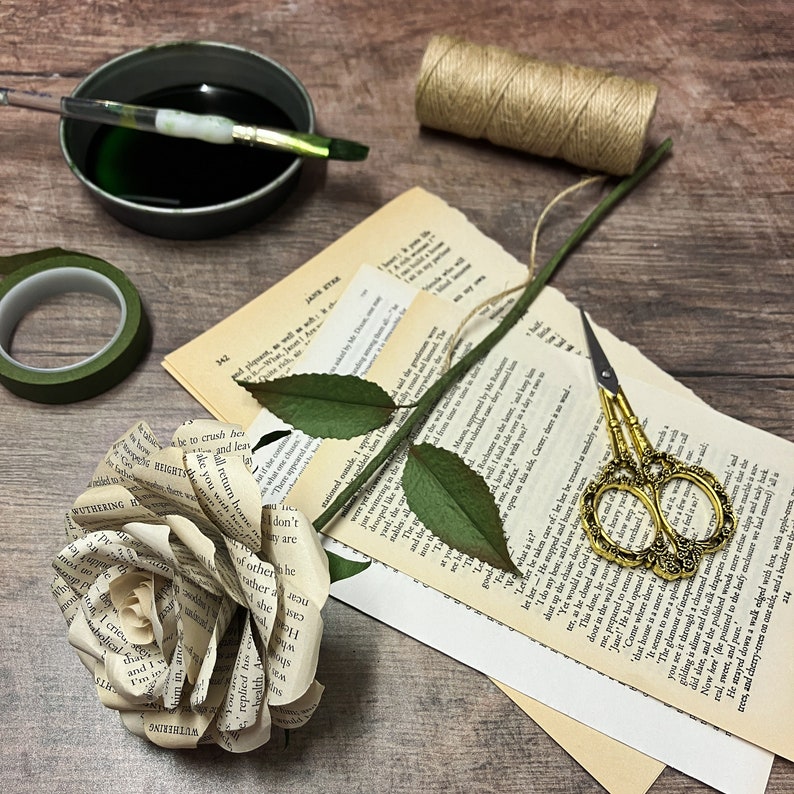May include: A paper rose crafted from the pages of a book, with a green stem and leaves, resting on a stack of books. The rose is surrounded by other crafting supplies, including a spool of twine, a paintbrush, and a jar of green paint.