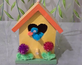 May include: A yellow birdhouse with an orange roof and a heart-shaped opening. The birdhouse has a blue bird inside and two green bushes with red and purple pom-poms.