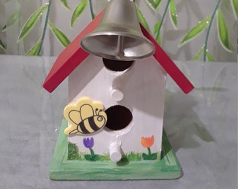 May include: A small wooden birdhouse painted white with a red roof and a bell on top. The birdhouse has a bee painted on the side and flowers painted on the bottom.