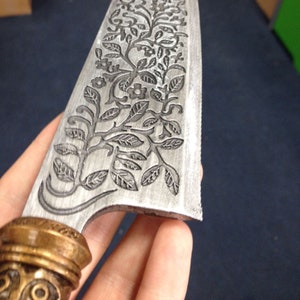 Puede incluir: Primer plano de una hoja de cuchillo de acero inoxidable con un diseño floral grabado. La hoja presenta un patrón detallado de hojas, flores y vides. El mango es de color dorado con un diseño ornamentado.