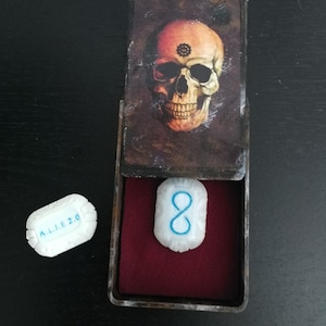 Puede incluir: Una pequeña caja de madera con un diseño de calavera en la tapa. La caja está abierta y contiene una baldosa de cerámica blanca con el número 8 inscrito en azul. Una segunda baldosa de cerámica blanca con el texto "A.L.I.E 2.0" es visible a la izquierda de la caja.