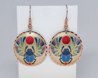 May include: A pair of gold-toned earrings with a round design featuring a blue scarab beetle with wings and a red sun. The earrings are made of copper and have a textured edge.