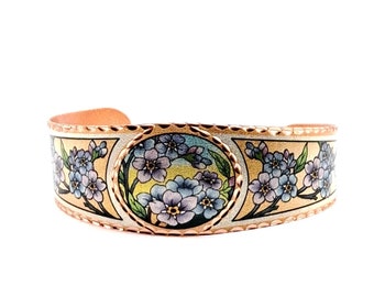 May include: A copper bracelet with a floral design. The bracelet is decorated with forget-me-not flowers in shades of blue, purple, and yellow. The flowers are surrounded by a gold-colored border.