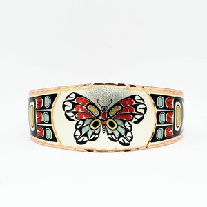 Puede incluir: Una pulsera de cobre con fondo blanco y un diseño de mariposa colorido. La mariposa tiene alas rojas, amarillas, azules y negras. La pulsera tiene una banda de cobre alrededor del borde.