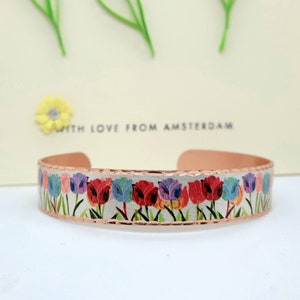 Puede incluir: Pulsera de cobre con fondo blanco y un estampado floral colorido de tulipanes. La pulsera tiene una textura martillada y un borde plateado. El texto "With Love From Amsterdam" es visible en el fondo.