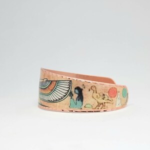 May include: Copper bracelet with an Egyptian-themed design. The design features a winged goddess, a bird, and other hieroglyphs. The bracelet is a warm copper color.