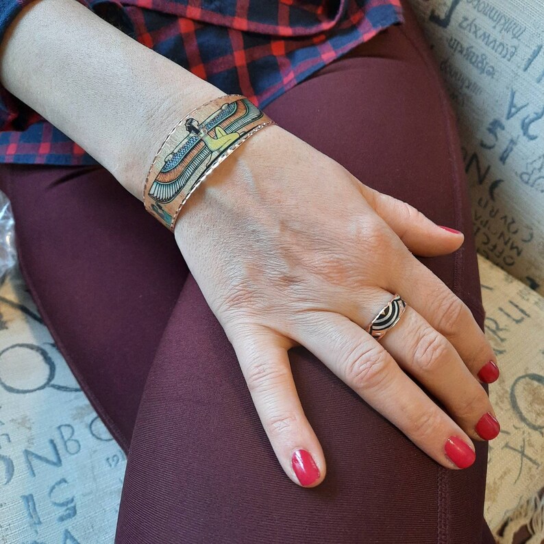 May include: A copper bracelet with a colorful image of an Egyptian goddess with wings. The bracelet is on a person's wrist. The person is wearing a red and black plaid shirt and maroon pants. The person is also wearing a ring with a black and white design on their finger.