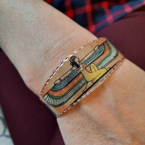 May include: A copper bracelet with a detailed image of an Egyptian goddess with wings. The goddess is depicted in a seated position with her arms outstretched.