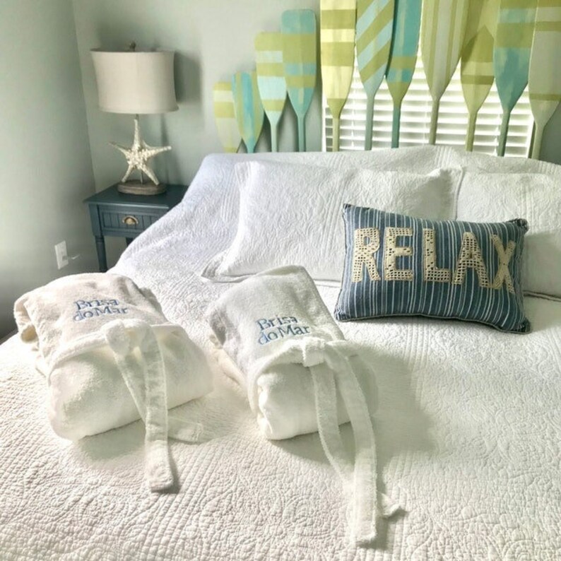 May include: Two white bathrobes with the text "Bris do Mar" embroidered on them are folded on a white bed with a blue and white pillow that says "Relax".
