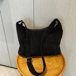 May include: A black corduroy shoulder bag with a long adjustable strap. The bag has a simple design and is made of a soft, textured fabric.