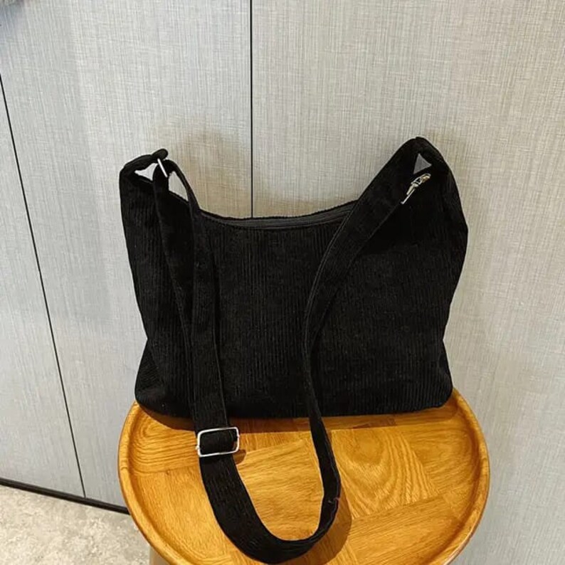 May include: A black corduroy shoulder bag with a long adjustable strap. The bag has a simple design and is made of a soft, textured fabric.