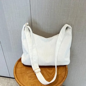 May include: A white corduroy shoulder bag with a long adjustable strap. The bag has a simple, minimalist design.