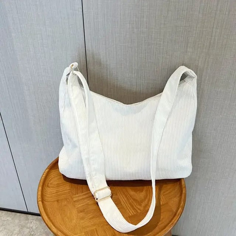 May include: A white corduroy shoulder bag with a long adjustable strap. The bag has a simple, minimalist design.