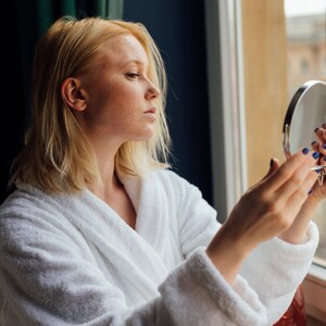 May include: A woman in a white bathrobe looks at her reflection in a round hand mirror. She is standing in front of a window with a view of a city.