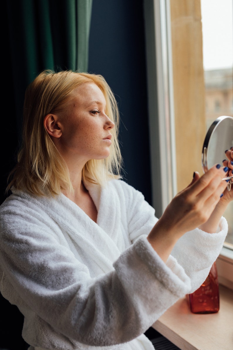 May include: A woman in a white bathrobe looks at her reflection in a round hand mirror. She is standing in front of a window with a view of a city.