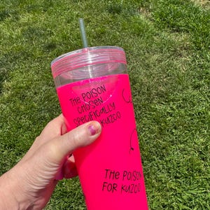 以下が含まれることがあります： 鮮やかなピンク色のラップが付いた透明なプラスチックタンブラー。ラップには、黒で「The Poison Chosen Specifically Kuzco」と「The Poison For Kuzco」の文字が書かれています。緑色のストローが蓋に挿入されています。タンブラーは緑の草を背景に持っています。