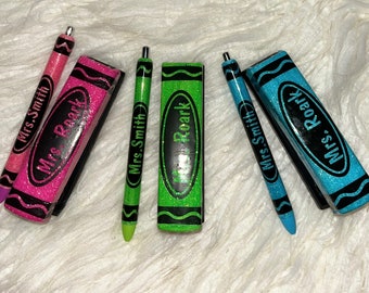 Puede incluir: Bolígrafos y gomas de borrar con forma de crayón brillantes en rosa, verde y azul. Los bolígrafos y las gomas de borrar tienen detalles en negro y los nombres "Mrs. Smith" y "Mrs. Roark" están escritos en ellos. Los artículos están diseñados para parecerse a crayones.
