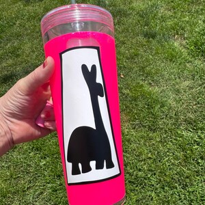 以下が含まれることがあります： 明るいピンク色のスリーブと蓋が付いた透明なプラスチックタンブラー。タンブラーには、ラマの白黒グラフィックが描かれています。緑色のストローが蓋に挿入されています。タンブラーは緑の草を背景に保持されています。