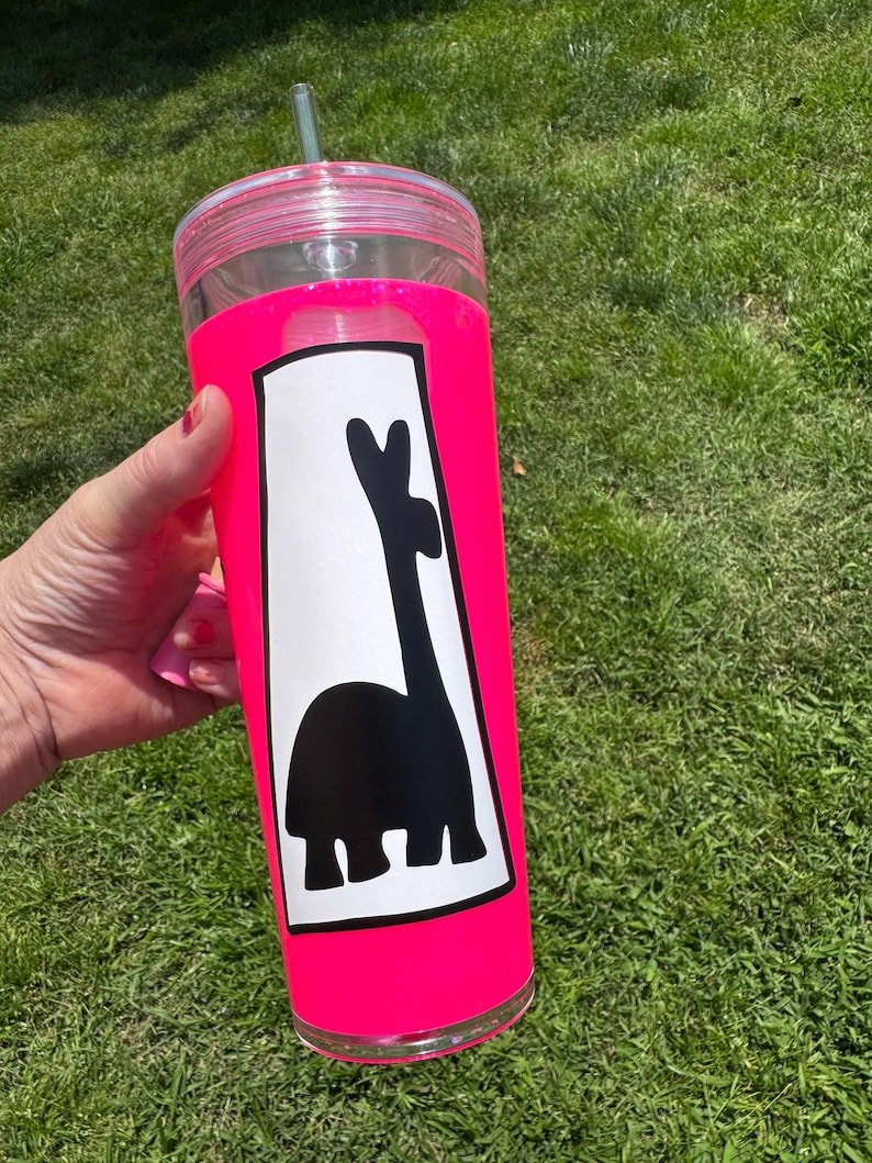 以下が含まれることがあります： 明るいピンク色のスリーブと蓋が付いた透明なプラスチックタンブラー。タンブラーには、ラマの白黒グラフィックが描かれています。緑色のストローが蓋に挿入されています。タンブラーは緑の草を背景に保持されています。