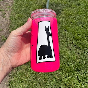 Puede incluir: Un vaso rosa brillante con purpurina y un diseño de llama negra sobre fondo blanco. Tiene una tapa transparente y una pajita verde. El vaso se sostiene en una mano, sobre un fondo de hierba verde.